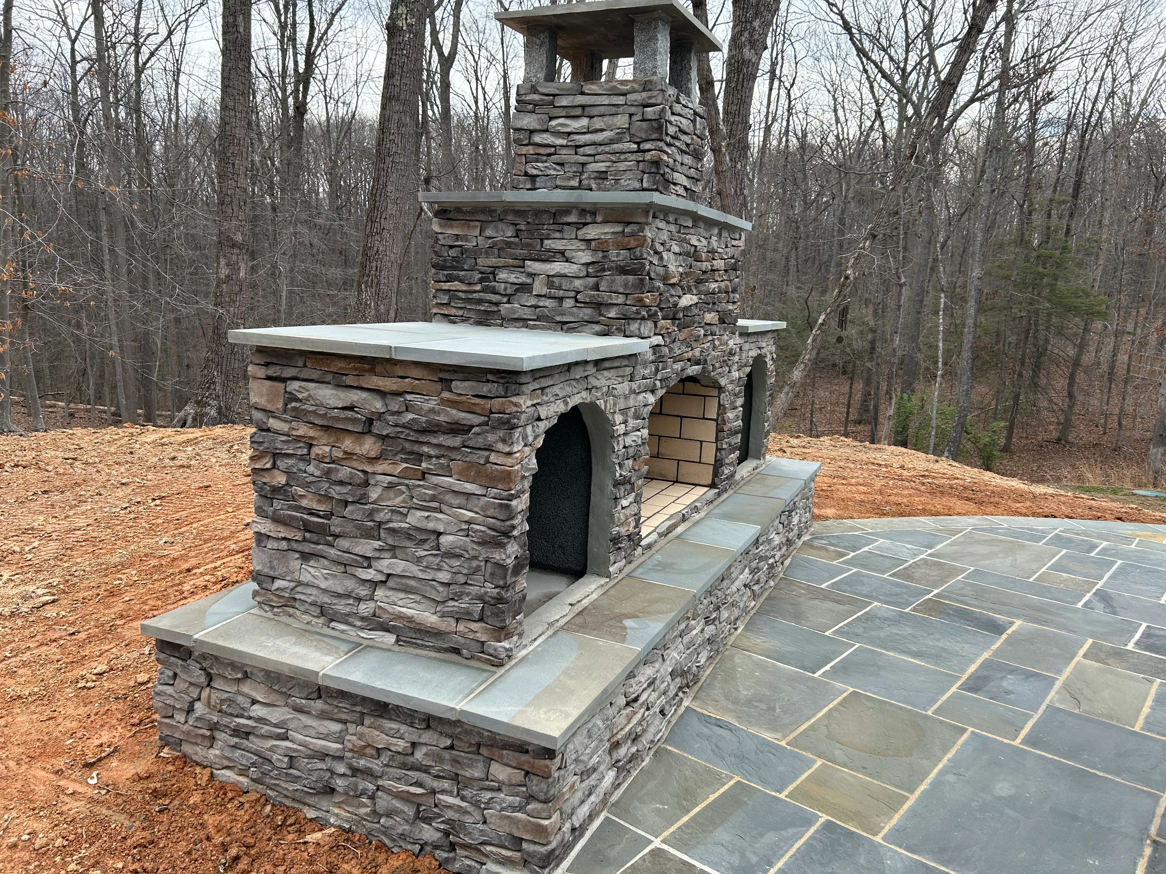 Stacked stone outdoor fireplace with chimney and flagstone patio in wooded backyard