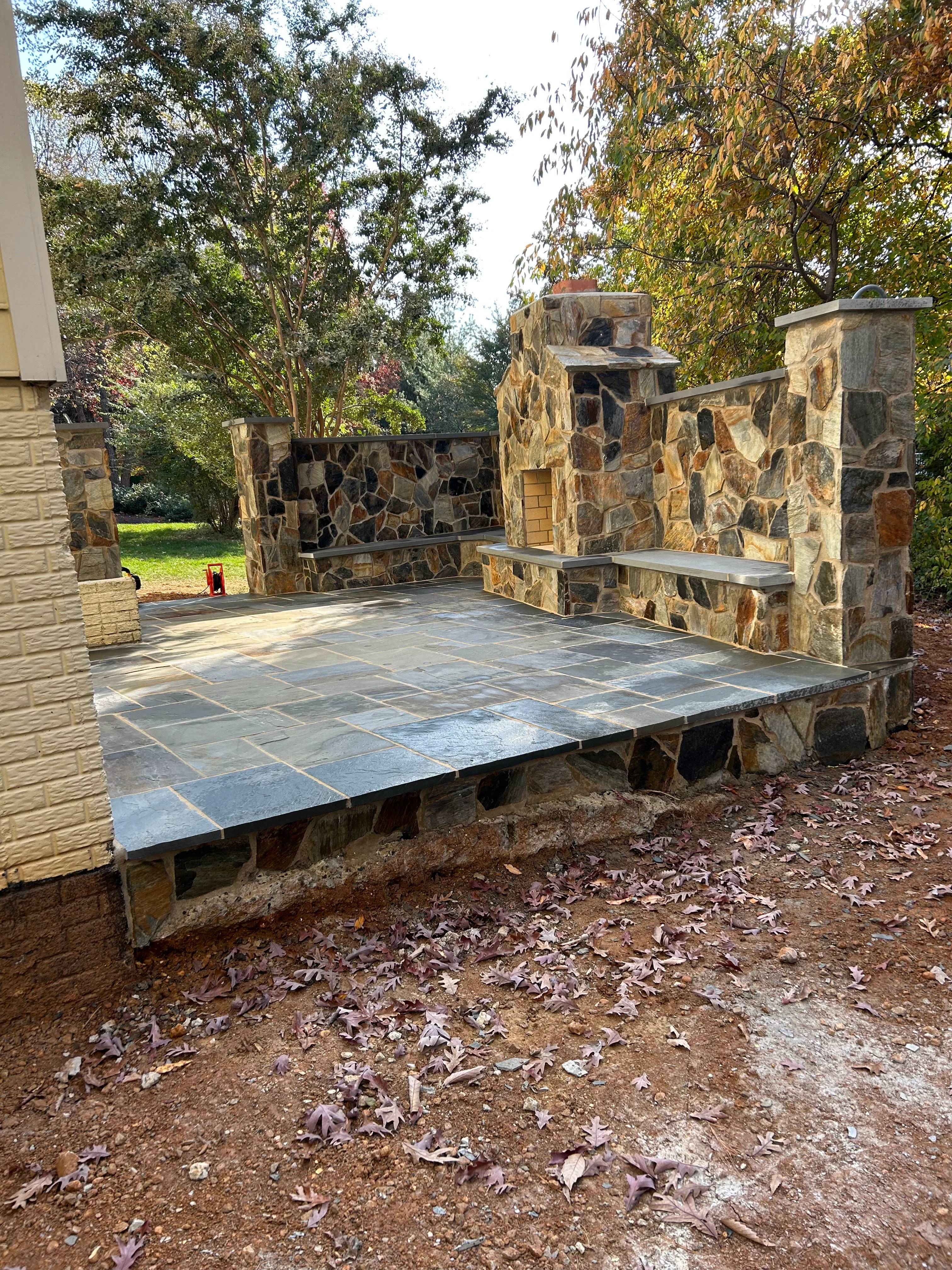 Stone patio with fireplace feature and built-in seating walls in wooded backyard