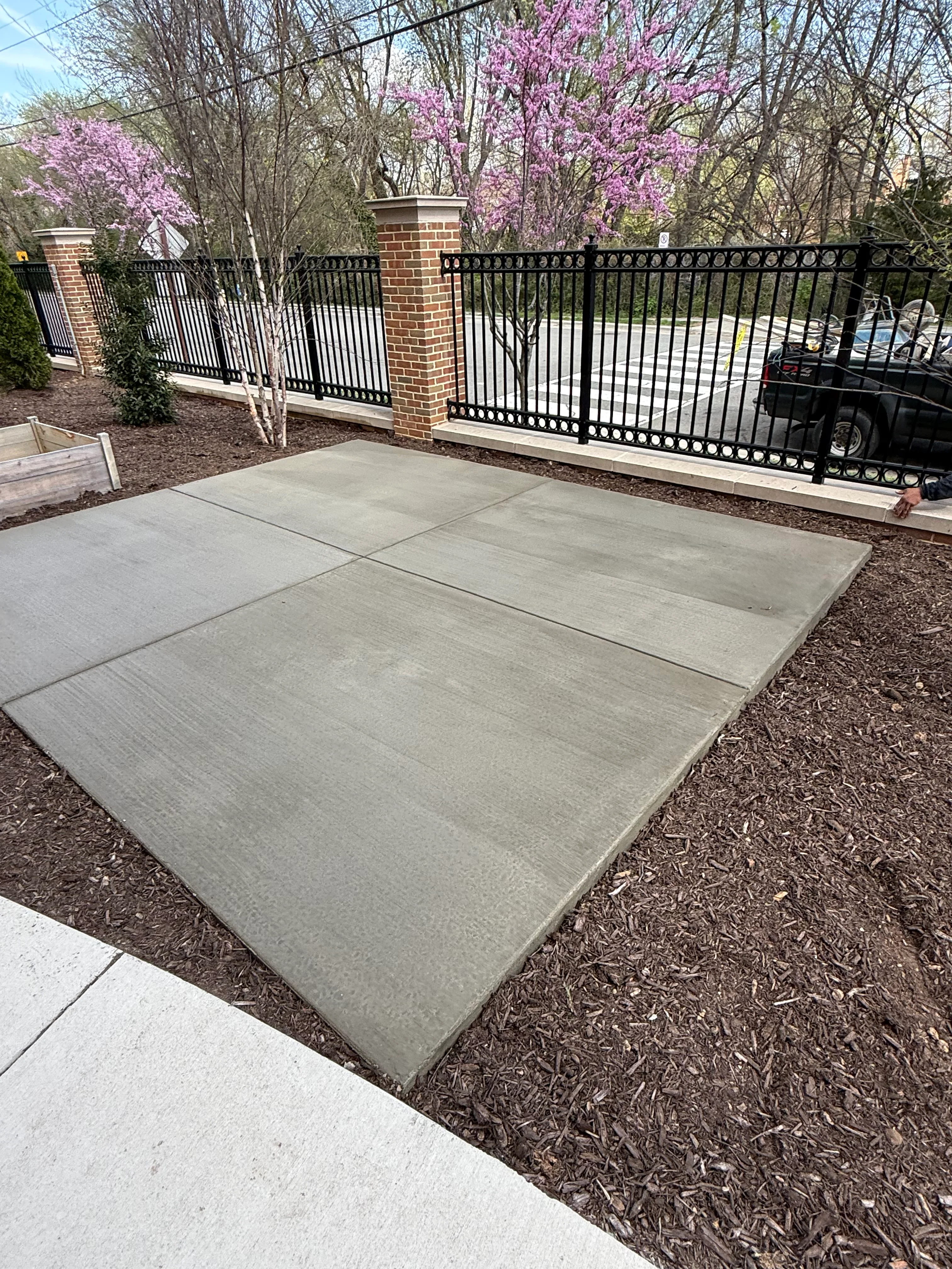 Gray concrete driveway with black iron gate and brick pillars in spring setting
