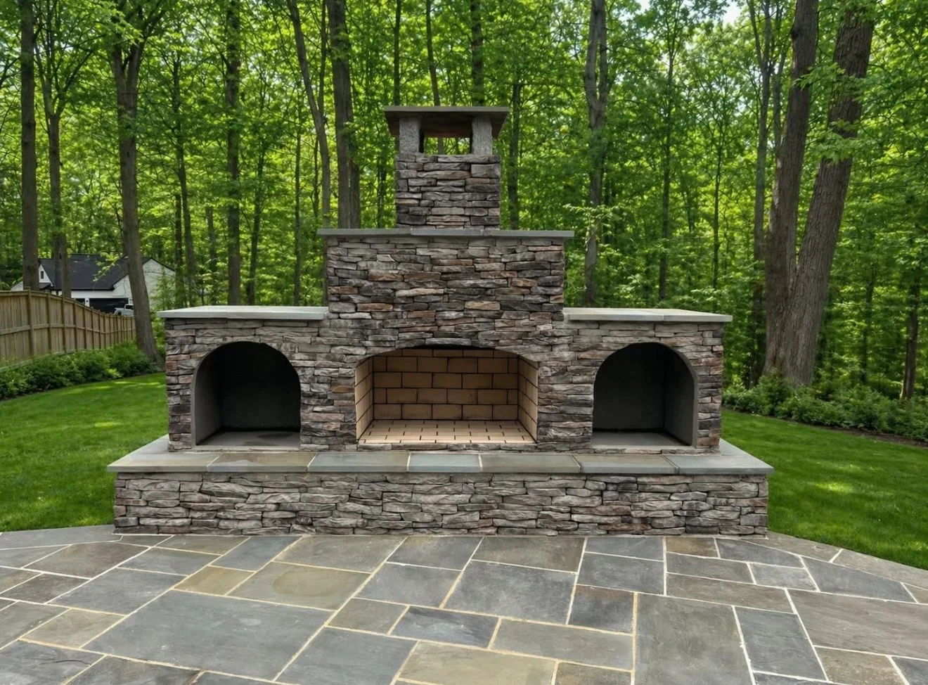 Stone fireplace with arched openings and chimney on flagstone patio surrounded by trees