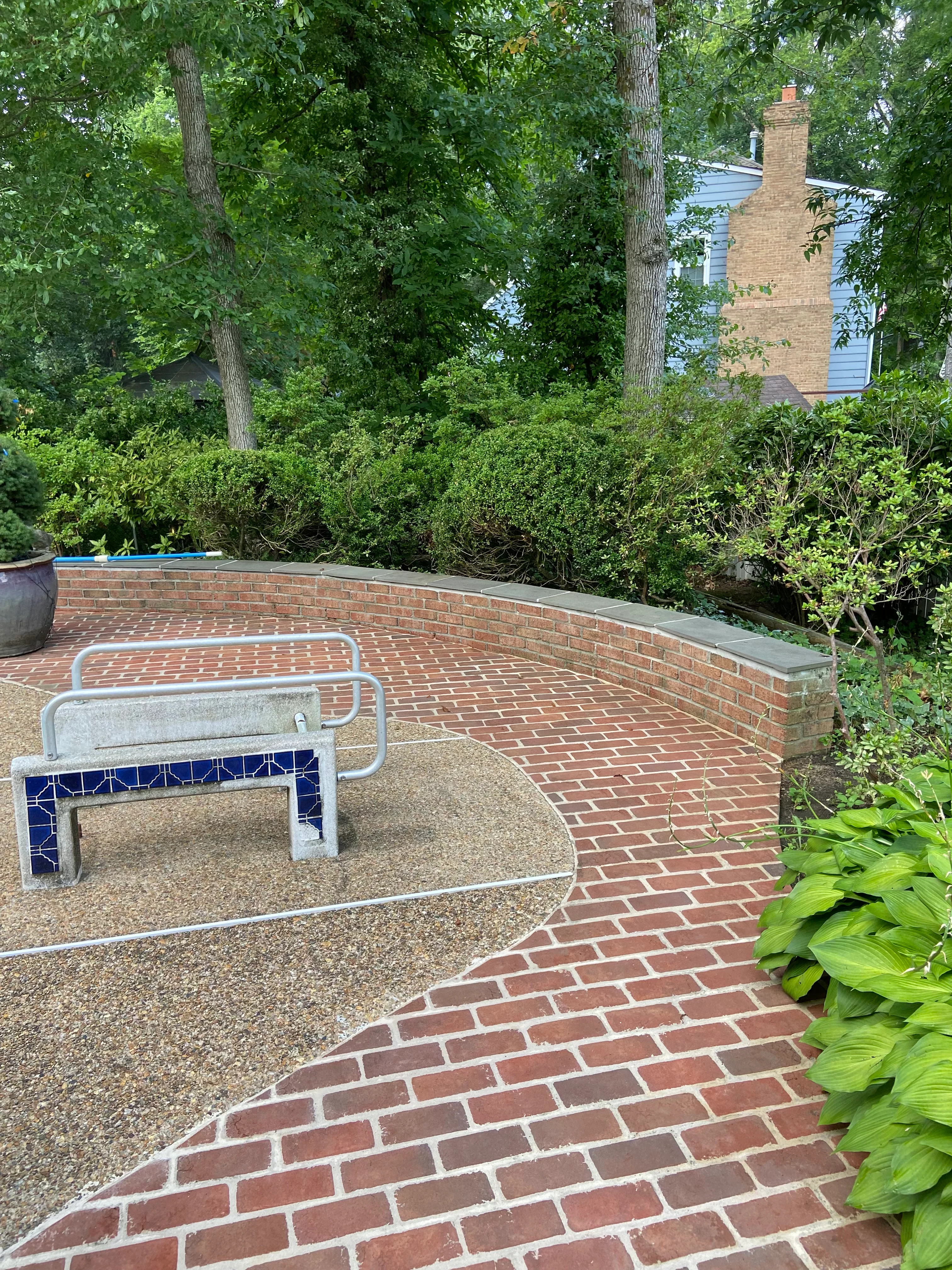 Brick patio with curved retaining wall and metal bench in wooded backyard