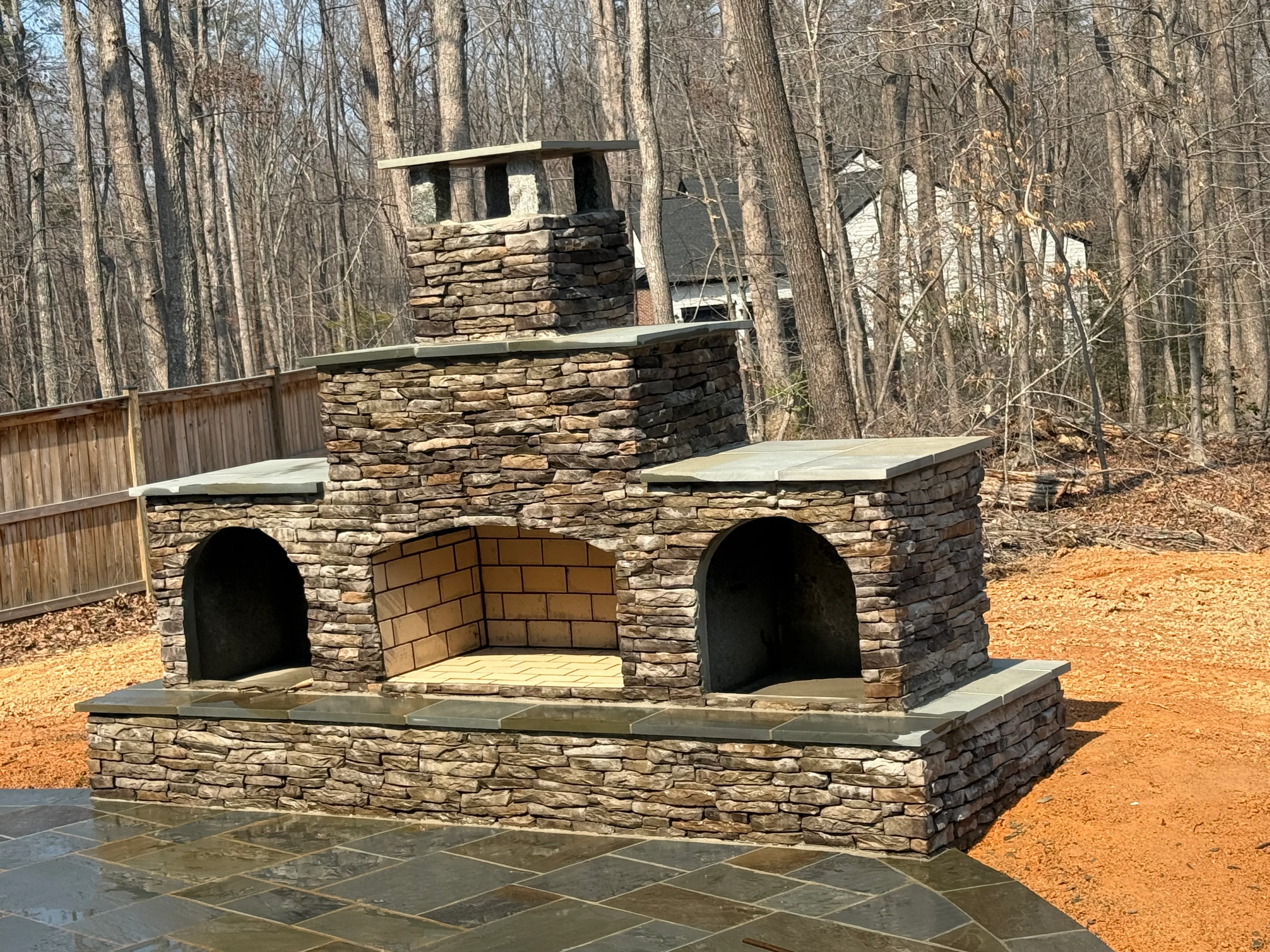 Stacked stone outdoor fireplace with brick firebox and chimney in wooded backyard