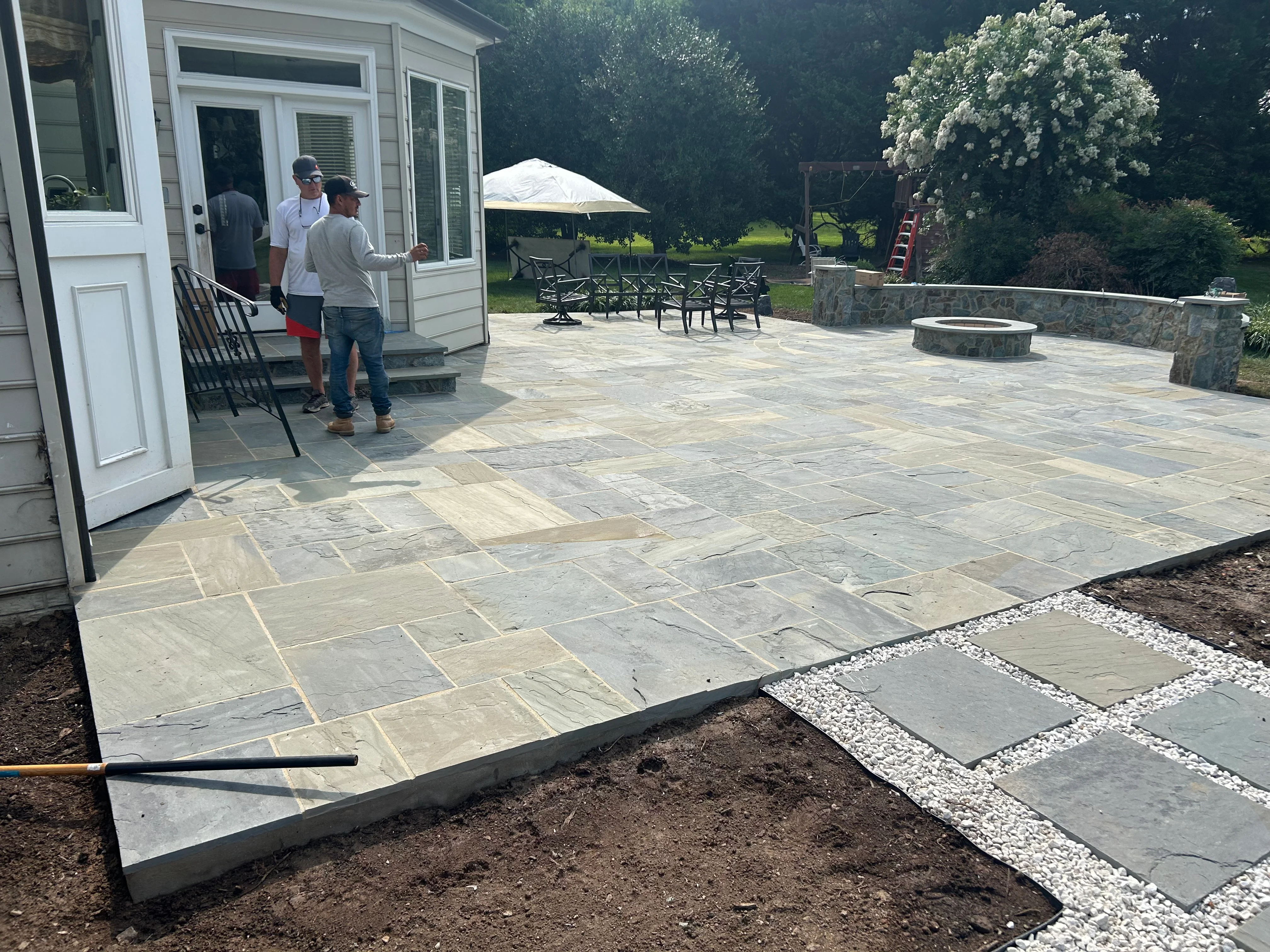 Large bluestone patio with fire pit, seating walls, and mature landscaping in residential backyard